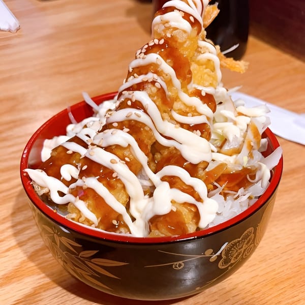 Crispy tempura shrimp with white and brown sauces, sesame seeds, and shredded cabbage in a red and black bowl.