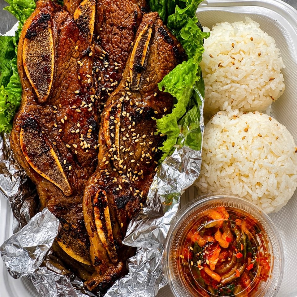 Grilled short ribs with sesame seeds, lettuce, two rice balls, and kimchi in a takeout container.